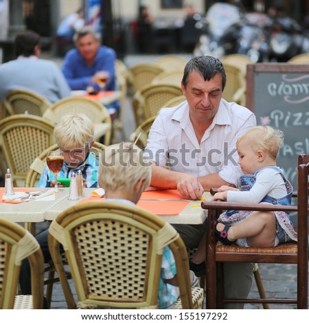 Family, a father with three children, two teenager twins sons, and little cute baby girl, having fun in cafe on summer terrace in a center of busy city street, boys playing with gadget smart phone