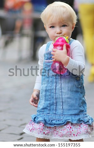Portrait of funny little baby girl walks on a busy street drinking water from plastic feeding bottle