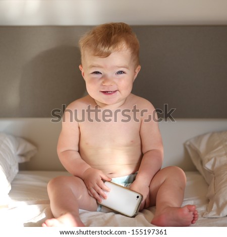Happy baby, just awake, sitting in parents bed playing with modern gadget mobile smart phone