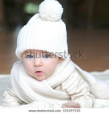 Portrait of cute little baby with big beautiful blue eyes laying on blanket wearing cozy warm white knitted hat and scarf