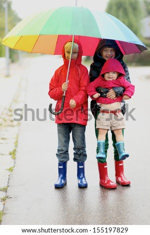 Three happy children, two teenager boys, twin brothers with little funny baby sister having fun walking together on the street sharing colorful umbrella on a rainy windy chilly autumn day