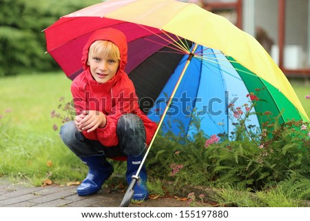 Cute kid, teenager boy in red rain coat and blue rubber boots hiding from rain sitting under colorful umbrella in front of the house