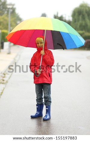 Cute kid, teenager boy in red rain coat and blue rubber boots walking on the street under colorful umbrella on a rainy windy chilly autumn day