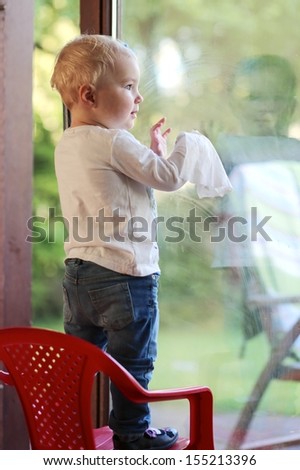 Funny little blond baby girl cleaning big sliding window door with wet serviette standing on small red plastic chair
