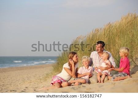 Happy family of five, mother, father, twin brothers and baby daughter are enjoying sunset at a beautiful beach sitting together in dunes next to the sea