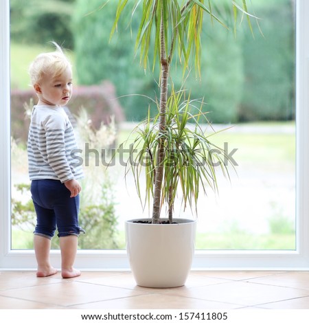 Beautiful Little Baby Girl With Blond Curly Hair Plays Inside Of The House Standing Next To A Big Window With Street View