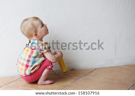 Cute little baby girl sitting on the floor inside of the house during repair works holding painting roll in her hands