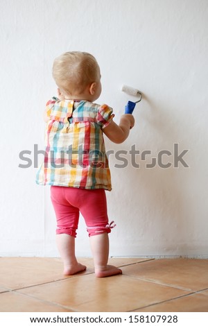 Funny little baby girl plays indoors during repair work in the house paining wall holding roll in her hands