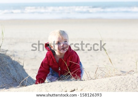 Sweet smiling little baby girl in a warm red coat crawls to the dunes along peaceful long sandy beach on a sunny autumn day