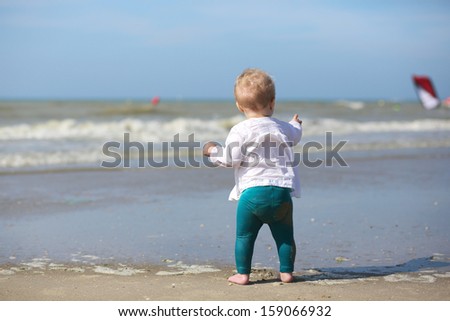 Cute blond baby girl in white blouse is standing next the water on the beach at a North Sea in Belgian coast on a sunny summer day looking at the yachts