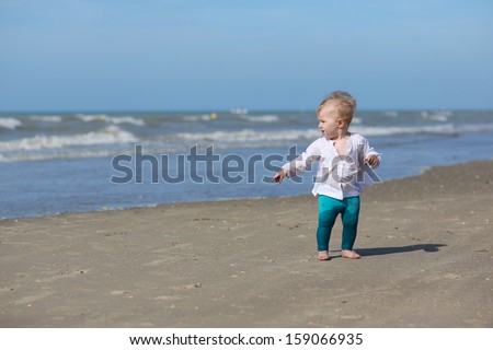Cute blond baby girl in white blouse is walking along the water on the beach at a North Sea in Belgian coast on a sunny summer day