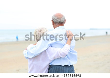 Portrait, back view of caring couple of two seniors, a man and his wife, standing together on a sandy peaceful beach looking at the ocean