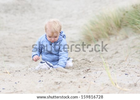 Lovely little baby girl in warm coat plays with sand in the dunes