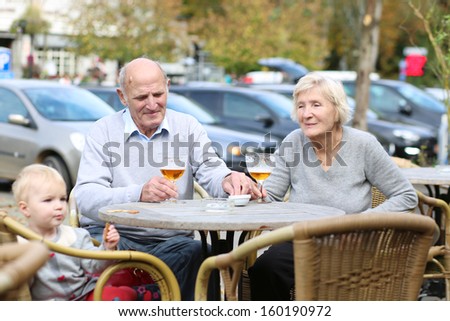 Happy smiling couple of seniors, a man and his wife, are enjoying glass of cold beer sitting with their granddaughter at the open air terrace of cozy cafe in the city center on a warm sunny day