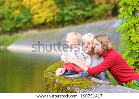 A happy family of three generations, mother, daughter, grandmother and little baby granddaughter, are standing together on the bridge of the river with the colorful trees around