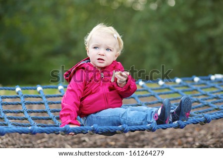 Funny little baby girl in a pink hooded warm coat relaxing on a swing net at playground in city park