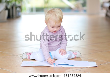 Funny lovely baby girl plays indoors drawing with colorful pencils sitting on tiles floor