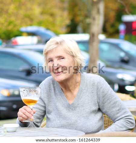Happy joyful senior woman sitting outdoors in cafe drinking cold fresh beer