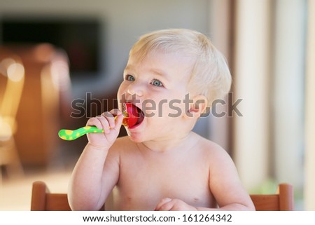 Adorable healthy baby with blond hair and blue eyes opens her mouth to eat fresh tomato holding a fork in her hand
