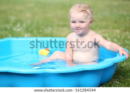 Funny little toddler splashing in small plastic baby bath outdoors in the garden on a hot summer day