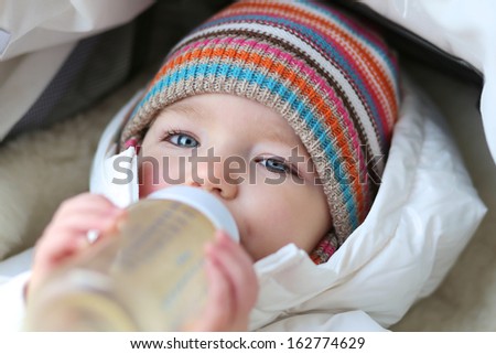 Little toddler girl laying cozy in white warm stroller drinking baby formula milk from the bottle