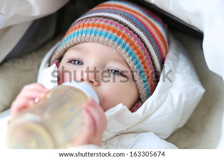 Cute little toddler girl laying cozy in white warm stroller drinking baby formula milk from the bottle