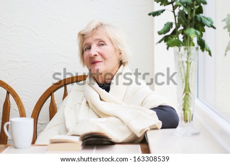 Happy healthy senior woman sitting comfortable in the kitchen wrapped in warm plaid reading interesting book and drinking delicious coffee on a cold rainy autumn day