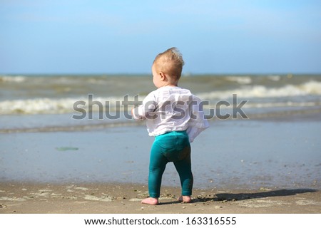 Cute blond baby girl in white blouse is standing next the water on the beach on a sunny summer day looking at the see