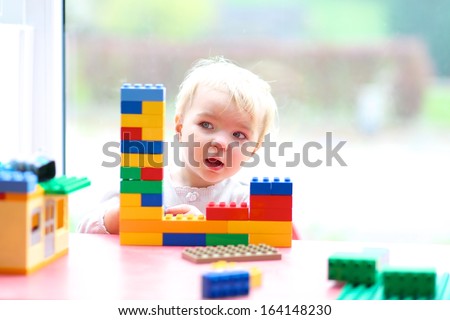 Cheerful Toddler Girl Building House From Plastic Blocks Sitting Next To A Big Window
