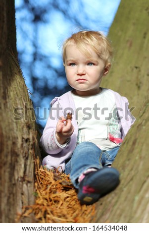 Funny little baby girl sitting cozy between the branches of the big old tree in the forest