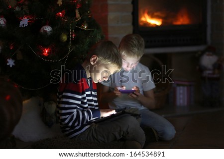 Two happy brothers sitting at the fireplace on a cozy winter evening nearby the christmas tree choosing gifts from online catalog or playing with tablet pc and smart phone