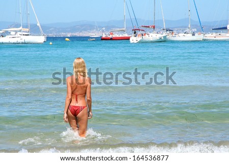 Beautiful young blonde healthy woman walking into the sea, looking at white yacht boats at the horizon