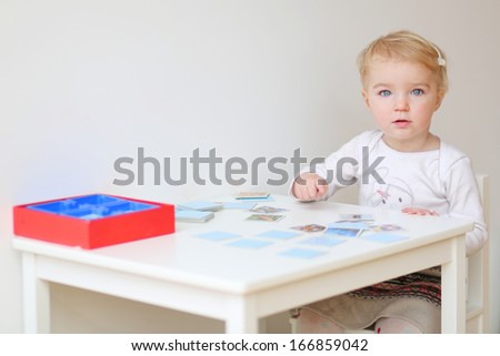 Cute blonde toddler girl playing memory game sitting at little white table indoors at home or kindergarten