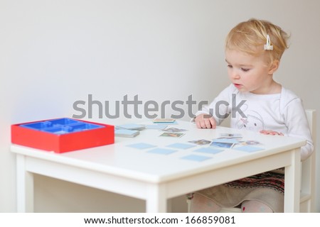 Adorable blonde toddler girl playing memory game sitting at little white table indoors at home or kindergarten