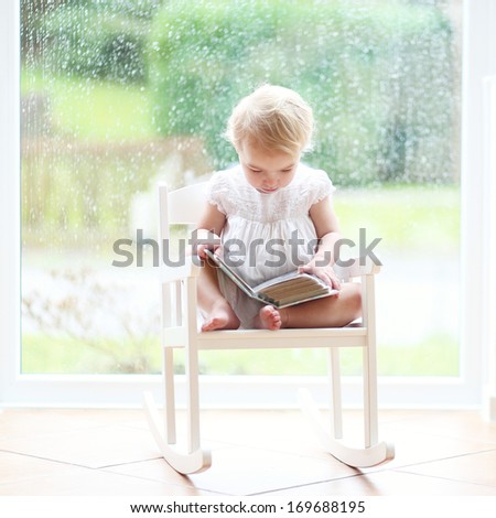 Sweet little blonde toddler girl sitting cozy in a white rocking chair next to a big window on a rainy day looking pictures in old family photo album