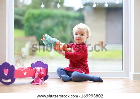 Sweet little blonde toddler girl playing with doll sitting on the tiles floor next to a big window