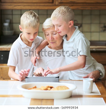 Teenager boys twin brothers teaching grandmother to play on tablet pc, enjoying time together, eating cookies and drinking coffee