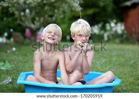 Two Cute Boys, Twin Brothers, Playing With Water In Little Plastic Bath Outdoors In The Garden At The Backyard Of The House