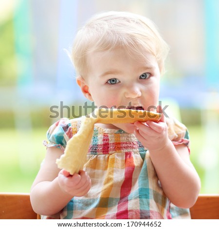 Cute blonde toddler girl eating tasty bread with butter sitting in the kitchen nearby big window with garden view