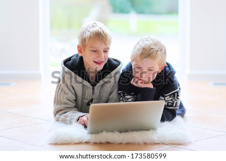 Two happy school boys, twin brothers, playing together with notebook laying indoor on the tiles floor