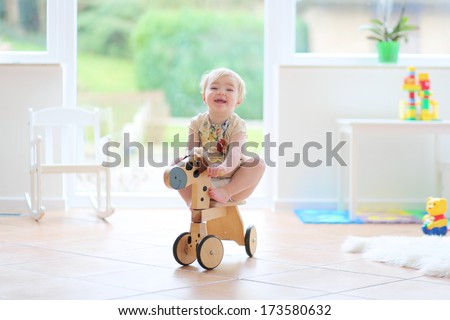 Happy little blonde toddler girl riding on wooden horse indoors in a beautiful room with big window and tiles floor
