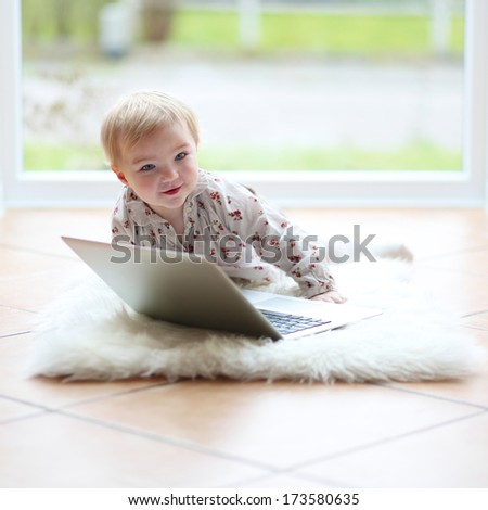 Cute blonde toddler girl playing with notebook sitting indoors on the tiles floor