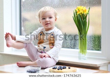 Funny blonde toddler girl enjoying pancakes sitting on the kitchen table next to a big window with narcissus flowers on a rainy spring day