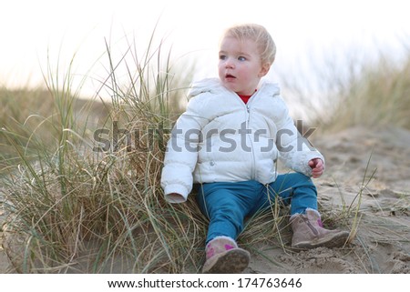 Adorable little toddler girl in warm white coat plays on a sunny early spring day with sand hiding in the dunes next to the beach in North Sea, Belgium, De Panne