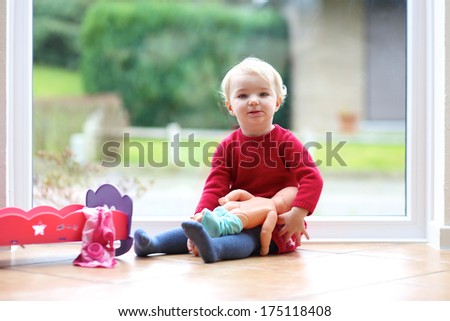 Lovely little blonde toddler girl playing with doll sitting on the tiles floor next to a big window