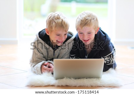 Two happy school boys, twin brothers, playing together with notebook laying indoor on the tiles floor