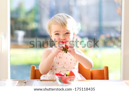 Cheerful blonde toddler girl in summer dress eating strawberries sitting indoors in the sunny kitchen next to a big window with garden view
