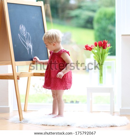 Cute blonde toddler girl in beautiful red dress drawing tulips with chalk on black board standing next to a big window