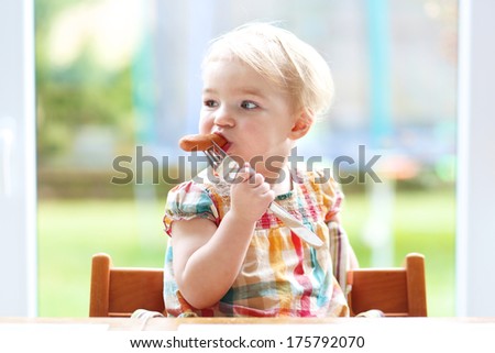 Cute blonde toddler girl eating biting on sausage from metal fork sitting in the kitchen nearby big window with garden view