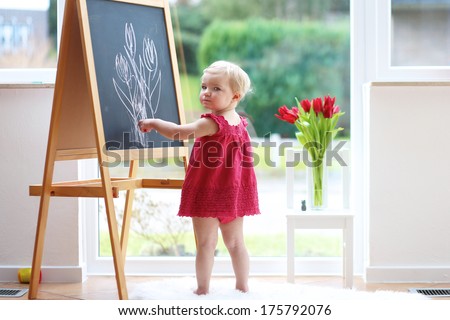 Adorable blonde toddler girl in beautiful red dress drawing tulips with chalk on black board standing next to a big window